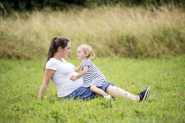 Pregnant woman sitting in grass with little daughter touching mother's belly - HAPF000719