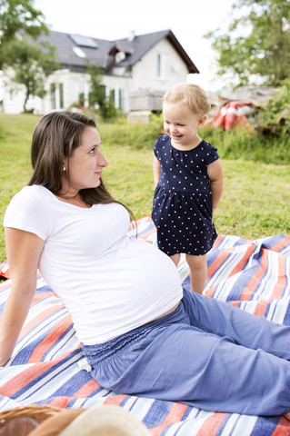 Schwangere Mutter sitzt mit ihrer kleinen Tochter auf einer Picknickdecke, lizenzfreies Stockfoto