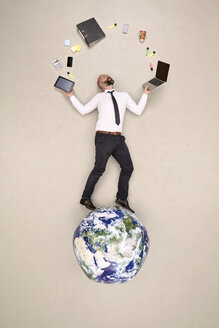 Businessman standing on globe juggling with office devices - BAEF001090