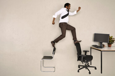 Businessman walking on chairs towards office desk - BAEF001228