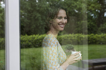 Smiling woman holding cup looking out of window - RBF004844
