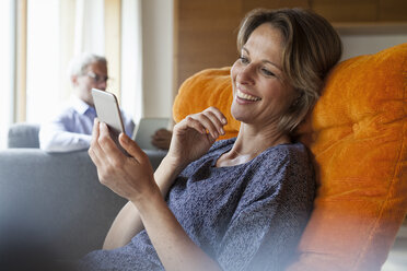 Lächelnde Frau zu Hause, die auf ihr Handy schaut, mit ihrem Mann im Hintergrund - RBF004902