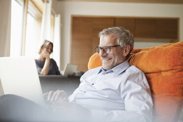 Smiling man using laptop in armchair with wife in background - RBF004905