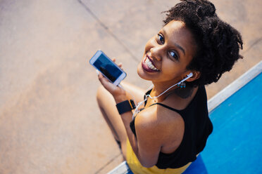 Lächelnde junge Frau mit Kopfhörern und Handy, die in einem Skatepark sitzt und in die Kamera blickt - GIOF001418
