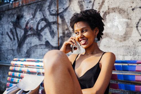 Porträt einer glücklichen jungen Frau, die auf einer Bank sitzt und mit einem Mobiltelefon telefoniert - GIOF001433
