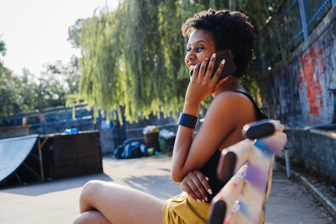 Happy young woman sitting on bench in a skatepark telephoning with cell phone stock photo