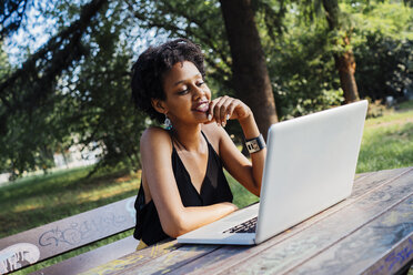 Junge Frau sitzt auf einer Parkbank und benutzt einen Laptop - GIOF001436