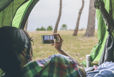 Rückenansicht einer jungen Frau mit Smartphone, die in einem Zelt liegt und von außen fotografiert - DAPF000259