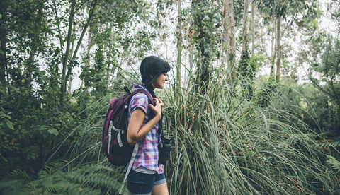 Lächelnde junge Frau mit Rucksack beim Spaziergang im Wald, lizenzfreies Stockfoto