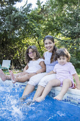 Portrait of smiling mother sitting on pool edge with her little daughters - ABZF000979