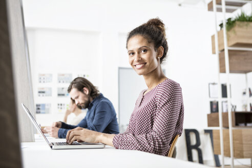 Lächelnde Frau im Büro bei der Arbeit am Laptop - RBF004914