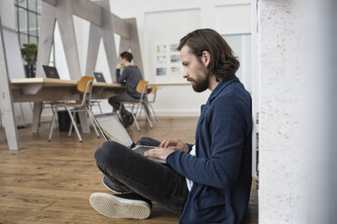 Mann sitzt auf dem Boden im Büro und arbeitet am Laptop - RBF004923