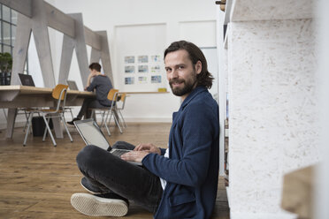 Lächelnder Mann sitzt auf dem Boden im Büro und arbeitet am Laptop - RBF004924