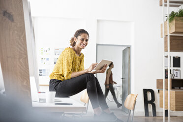 Lächelnde Frau sitzt auf einem Bürotisch und benutzt ein digitales Tablet - RBF004933