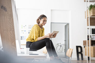 Lächelnde Frau sitzt auf einem Bürotisch und benutzt ein digitales Tablet - RBF004934
