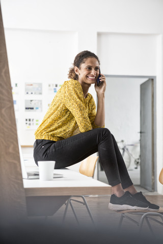 Lächelnde Frau sitzt auf einem Bürotisch und benutzt ein Mobiltelefon, lizenzfreies Stockfoto