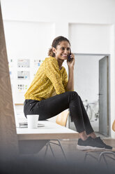 Lächelnde Frau sitzt auf einem Bürotisch und benutzt ein Mobiltelefon - RBF004935
