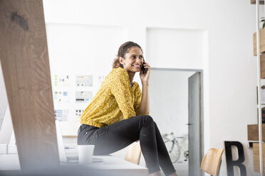 Lächelnde Frau sitzt auf einem Bürotisch und benutzt ein Mobiltelefon - RBF004937