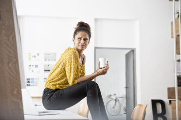 Lächelnde Frau sitzt auf einem Bürotisch und hält eine Tasse - RBF004938