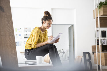 Smiling woman sitting on office desk working on paper - RBF004939