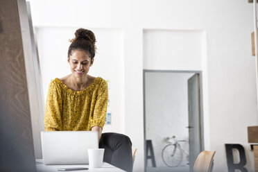 Smiling woman sitting on office desk using laptop - RBF004940