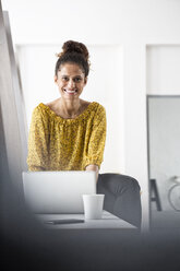 Lächelnde Frau sitzt auf einem Bürotisch und benutzt einen Laptop - RBF004941