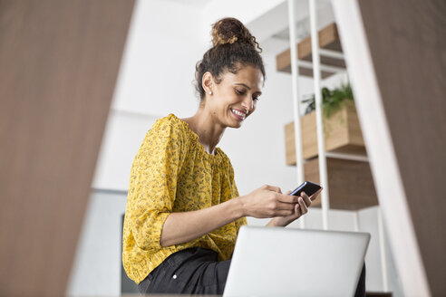 Lächelnde Frau sitzt auf einem Bürotisch und benutzt ein Mobiltelefon - RBF004944