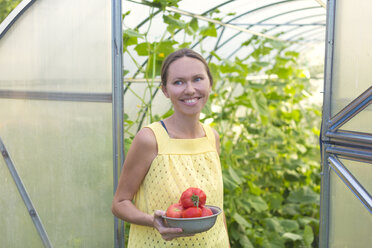 Happy woman with bowl of harvested tomatoes in front of greenhouse - KNTF000442