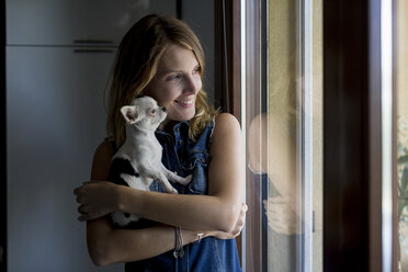 Lächelnde Frau mit Hund auf dem Arm schaut durch ein Fenster - MAUF000810