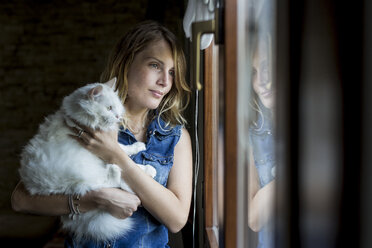 Smiling woman with cat on her arms looking through window - MAUF000820