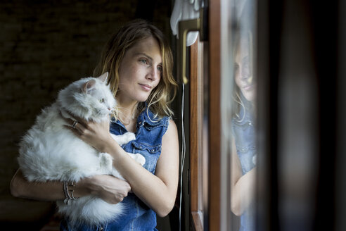 Lächelnde Frau mit Katze auf dem Arm schaut durch ein Fenster - MAUF000820
