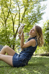 Glückliche Frau sitzt mit ihrem Chihuahua auf einer Wiese im Garten - MAUF000824
