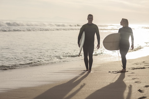 Paar mit Surfbrettern, das bei Sonnenuntergang am Strand spazieren geht, lizenzfreies Stockfoto