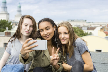 Germany, Berlin, three teenage girls sitting on roof top taking selfie with smartphone - OJF000146