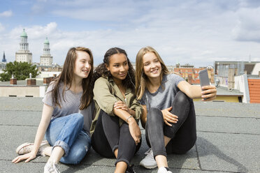 Germany, Berlin, three teenage girls sitting on roof top taking selfie with smartphone - OJF000147