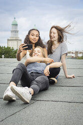 Deutschland, Berlin, drei Freunde sitzen auf dem Dach und machen ein Selfie mit Smartphone - OJF000164