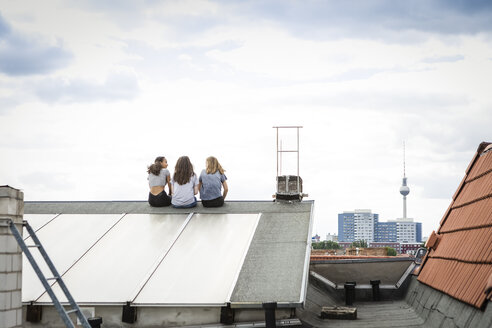 Germany, Berlin, back view of three friends sitting side by side on roof top - OJF000171