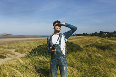 Relaxed young man with camera standing on a dune looking at distance - BOYF000540