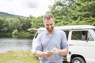 Smiling man using cell phone at a van at lakeside - FMKF002794