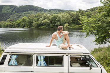 Man sitting on roof of a van at lakeside - FMKF002815