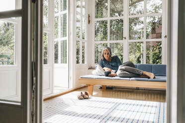 Woman lying on lounge in winter garden reading e-book - KNSF000252