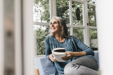 Lächelnde Frau mit Buch, die auf dem Sofa im Wintergarten sitzt und durch das Fenster schaut - KNSF000257