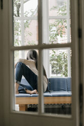 Rückenansicht einer Frau, die auf einem Sofa im Wintergarten sitzt und durch ein Fenster schaut - KNSF000263