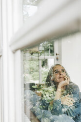 Lächelnde Frau sitzt hinter einer Fensterscheibe und schaut in die Ferne - KNSF000267