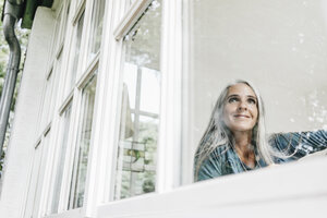 Lächelnde Frau zu Hause, die durch ein Fenster schaut - KNSF000268