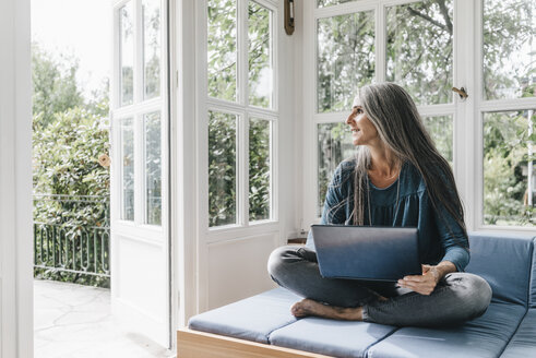 Woman with laptop sitting on lounge in winter garden looking through opened terrace door - KNSF000269