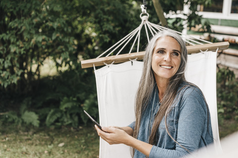 Porträt einer lächelnden Frau mit Smartphone, die in einer Hängematte im Garten sitzt, lizenzfreies Stockfoto