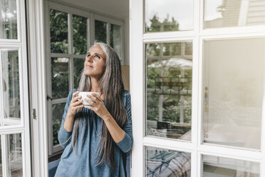 Lächelnde Frau lehnt am Türrahmen ihres Wintergartens und schaut mit einer Tasse Kaffee nach oben - KNSF000292
