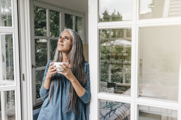 Smiling woman leaning at door frame of her winter garden with cup of coffee looking up - KNSF000292