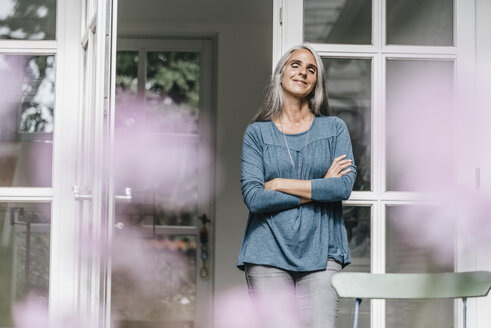 Smiling woman leaning at door frame of her winter garden enjoying sunlight - KNSF000313